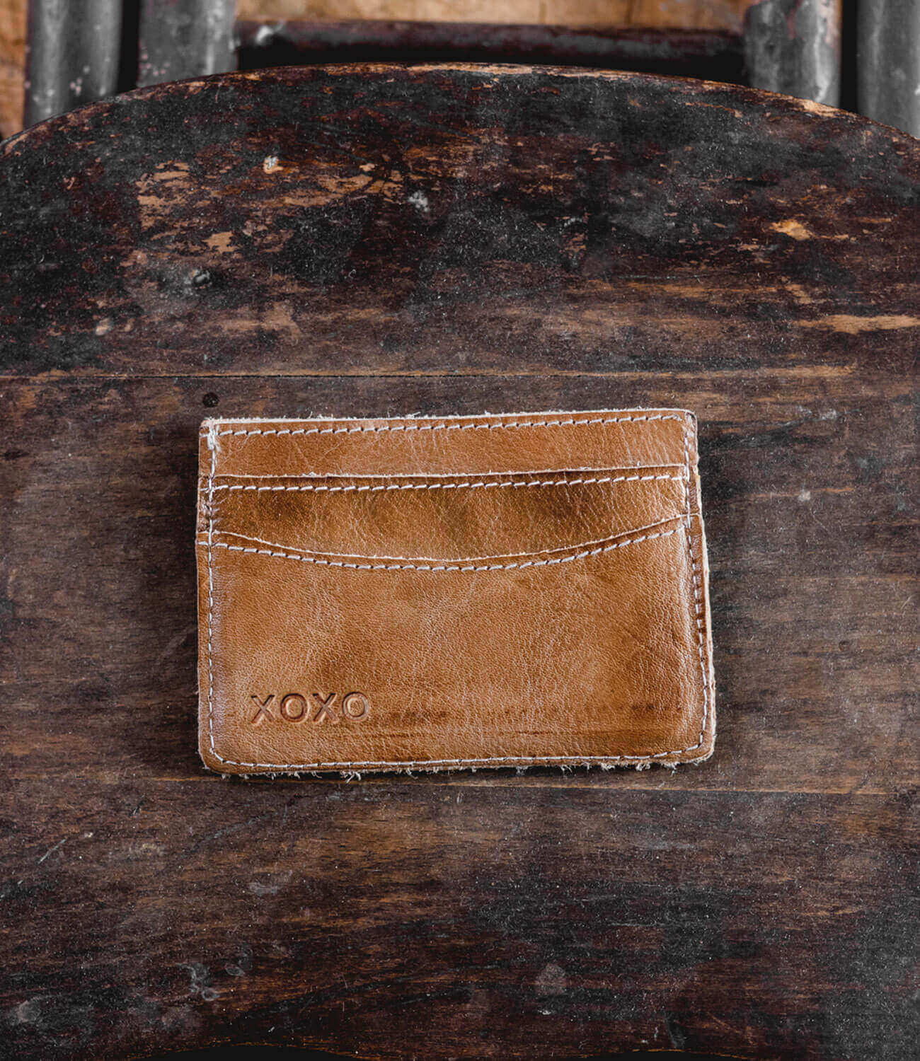 A brown leather compact card wallet named "Chuck" by Bed|Stü, featuring visible stitching and the word "XOXO" embossed in the corner, placed on a worn wooden surface—perfect for everyday carry.