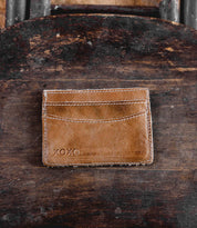 A brown leather compact card wallet named "Chuck" by Bed|Stü, featuring visible stitching and the word "XOXO" embossed in the corner, placed on a worn wooden surface—perfect for everyday carry.