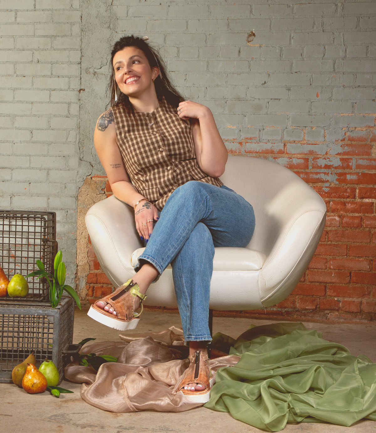 A woman in a sleeveless checkered top and jeans sits smiling in a white chair, surrounded by pears, fabric, and brick walls, her look completed with the Bed|Stü Clancy platform shoes featuring cushioned insoles for all-day comfort.