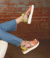 A person wearing the Bed|Stü Clancy metallic platform shoes—featuring green heel straps, white cushioned insoles, and leather cutout details—paired with blue jeans, feet elevated against a red brick wall background.