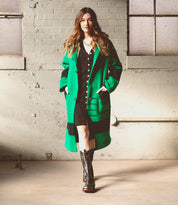 A woman stands indoors with her hands in her pockets, wearing a green coat over a black dress and striking red Bed|Stü Clarisse leather boots with a western heel, as sunlight streams through the windows behind her.