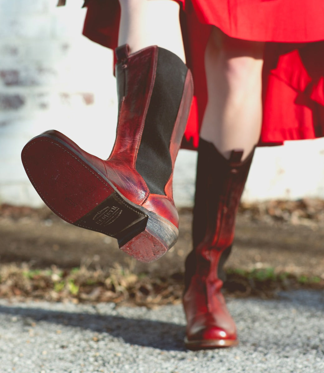 Model showcases the Clarisse mid-calf cowboy boots in bold red leather by Bed|Stü, pairing them with a matching red skirt; one boot is lifted to display the distinct western heel and sole, while the other remains grounded outdoors.