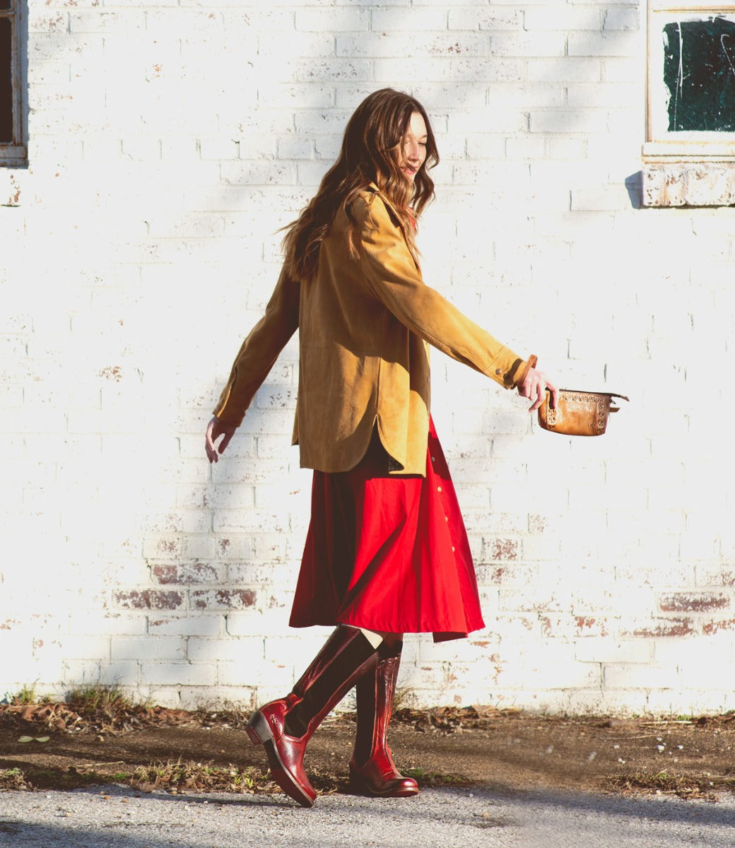 A person in a tan jacket, red skirt, and striking Bed|Stü Clarisse red leather boots with a western heel walks outside along a white brick wall, holding a small brown pot.