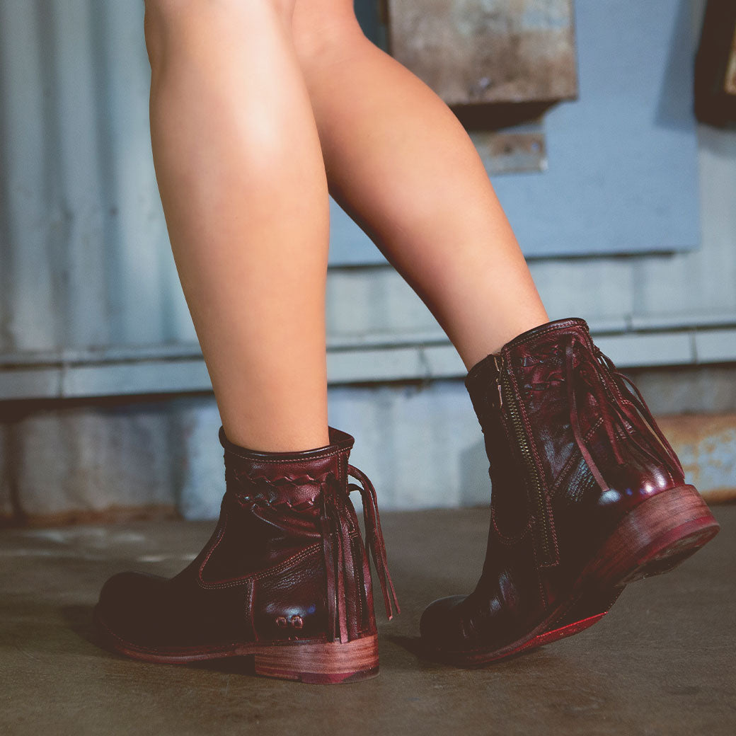 A person wearing the Bed|Stü Craven dark brown ankle boots featuring tassels and a hand-finished leather finish stands on a concrete floor in an industrial setting.