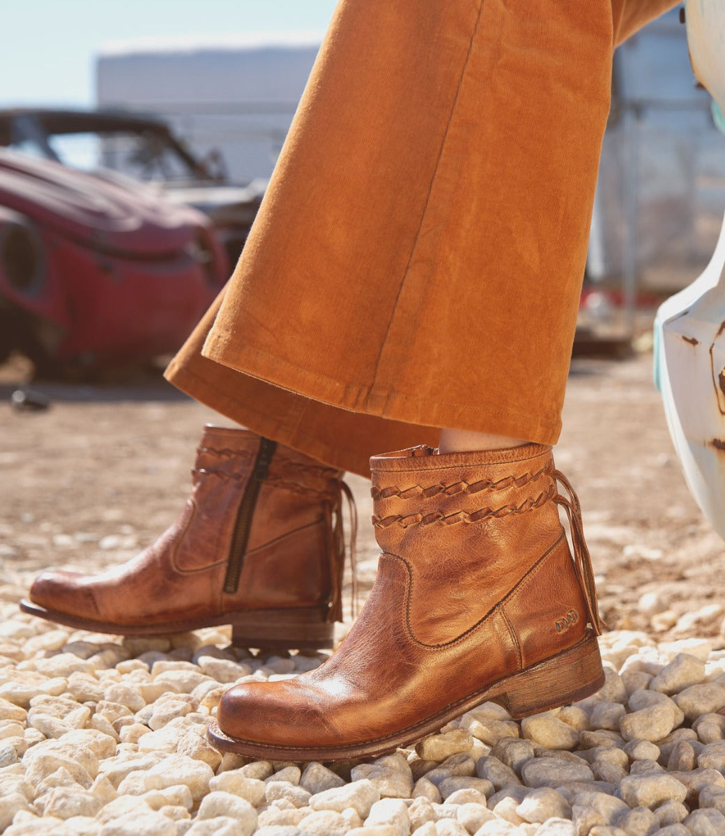 A person wearing the Bed|Stü Craven brown ankle boots with a woven shaft detail and rust-colored pants stands on light-colored gravel outdoors.