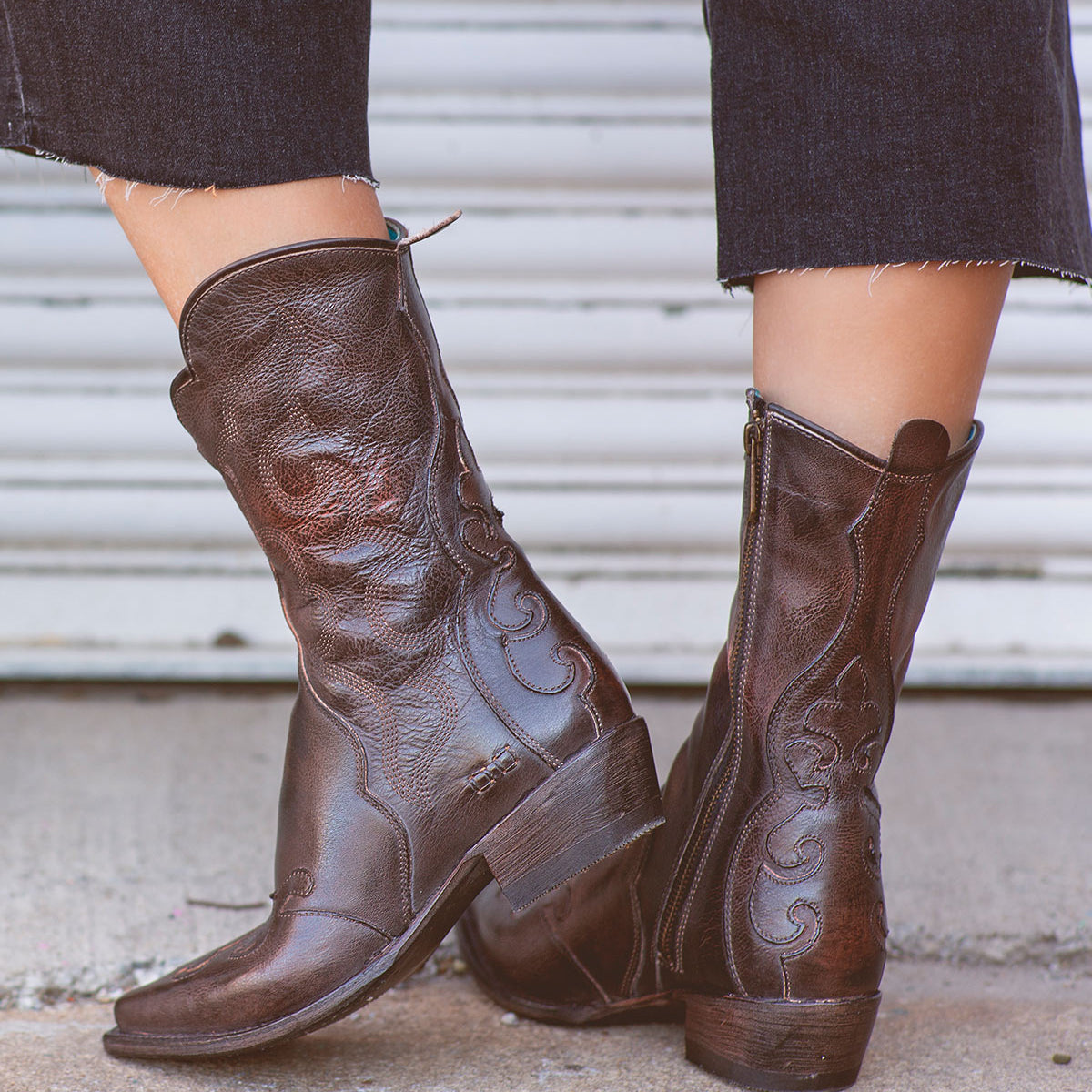 Wearing the Deuce boots by Bed|Stü—dark brown Western boots made from vegetable-tanned leather with intricate stitching and convenient side zippers—a person stands on a concrete surface in front of a metal door.