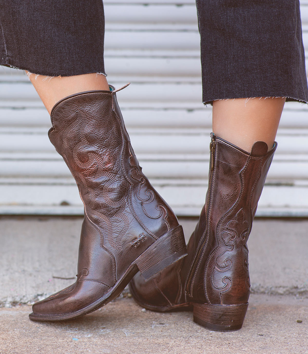 Wearing the Deuce boots by Bed|Stü—dark brown Western boots made from vegetable-tanned leather with intricate stitching and convenient side zippers—a person stands on a concrete surface in front of a metal door.