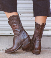 Wearing the Deuce boots by Bed|Stü—dark brown Western boots made from vegetable-tanned leather with intricate stitching and convenient side zippers—a person stands on a concrete surface in front of a metal door.