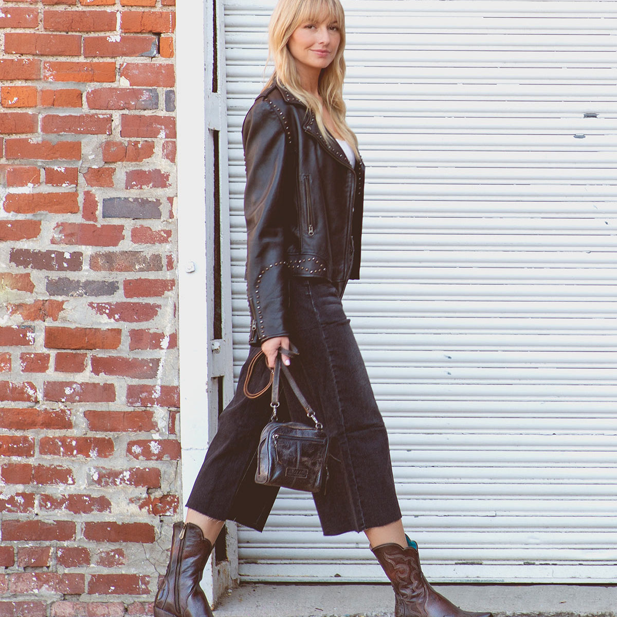 A woman in Bed|Stü's Deuce Western boots with detailed stitching steps outside, pairing them with a black leather jacket, cropped black jeans, and a small handbag. She walks past a brick wall and white metal garage door.