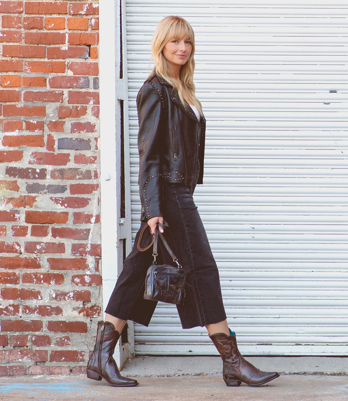 A woman in Bed|Stü's Deuce Western boots with detailed stitching steps outside, pairing them with a black leather jacket, cropped black jeans, and a small handbag. She walks past a brick wall and white metal garage door.