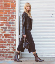 A woman in Bed|Stü's Deuce Western boots with detailed stitching steps outside, pairing them with a black leather jacket, cropped black jeans, and a small handbag. She walks past a brick wall and white metal garage door.