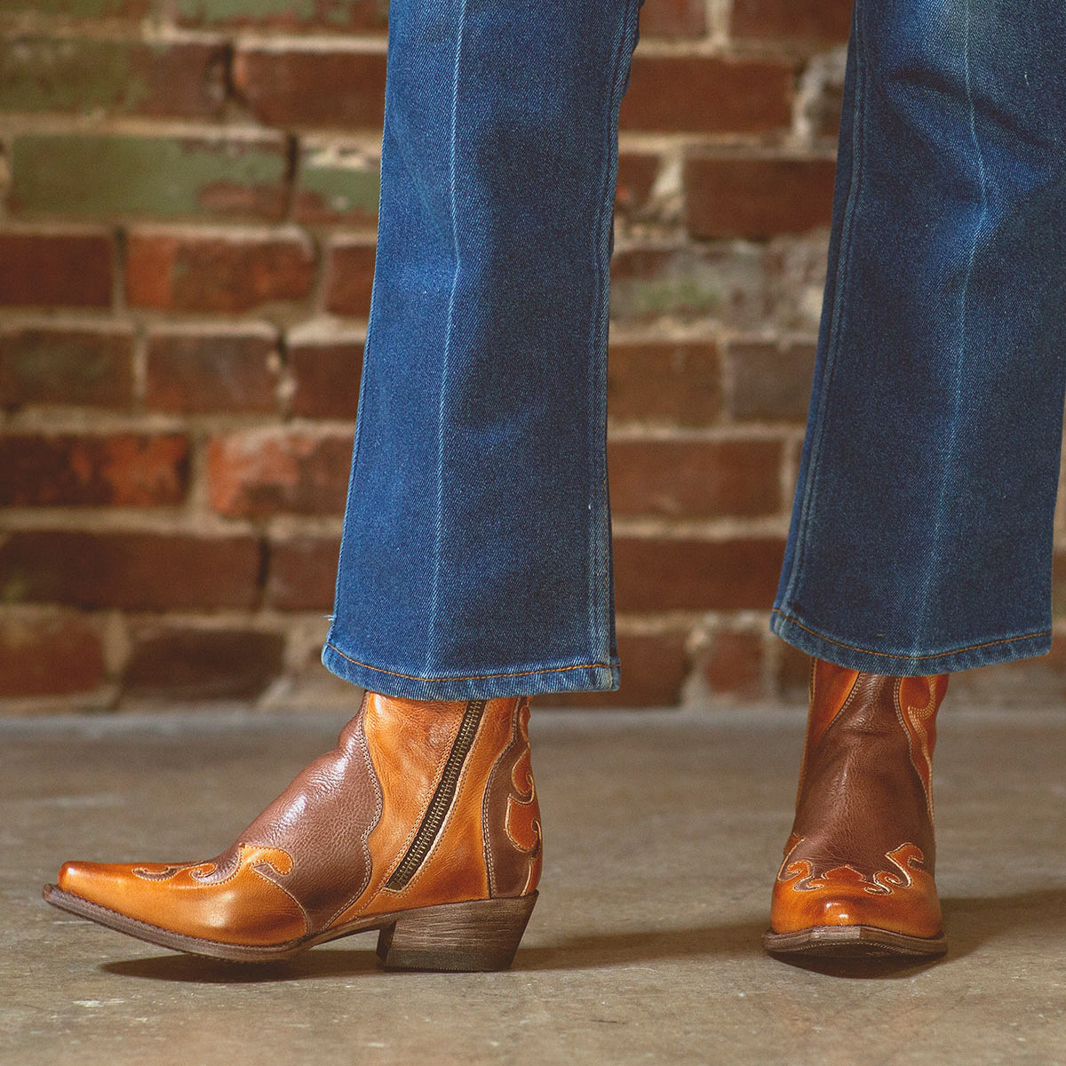 A person is wearing Bed|Stü Deuce brown leather Western boots with detailed stitching, paired with blue jeans, standing on a concrete floor in front of a brick wall.