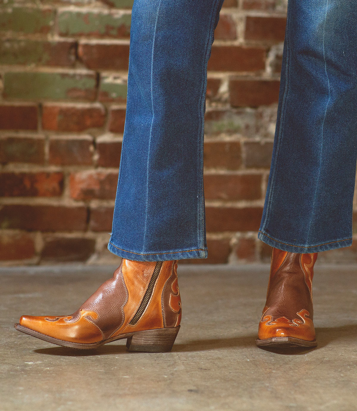 A person is wearing Bed|Stü Deuce brown leather Western boots with detailed stitching, paired with blue jeans, standing on a concrete floor in front of a brick wall.