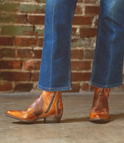 A person is wearing Bed|Stü Deuce brown leather Western boots with detailed stitching, paired with blue jeans, standing on a concrete floor in front of a brick wall.