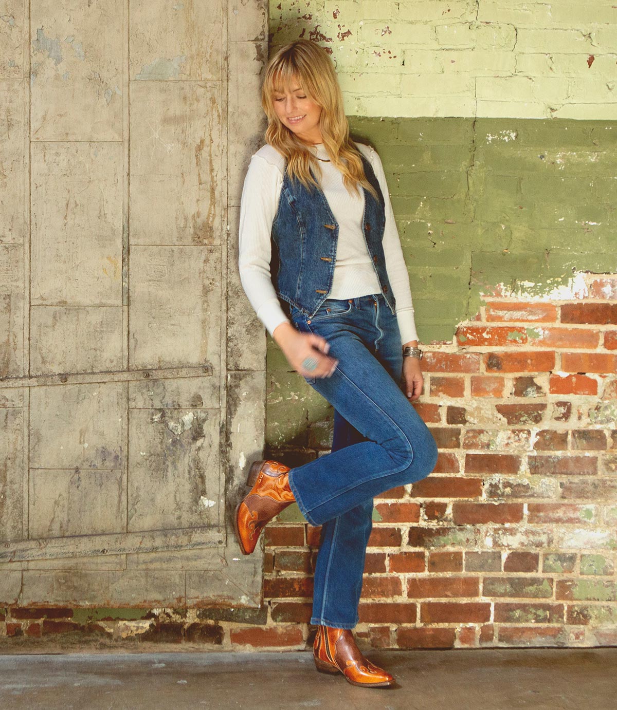A person in a white shirt, denim vest, and jeans stands against a weathered wall with one leg crossed, looking down and smiling, wearing the Deuce by Bed|Stü—shiny Western boots crafted from vegetable-tanned leather.