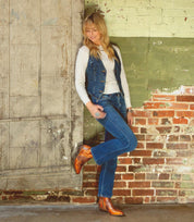 A person in a white shirt, denim vest, and jeans stands against a weathered wall with one leg crossed, looking down and smiling, wearing the Deuce by Bed|Stü—shiny Western boots crafted from vegetable-tanned leather.