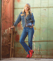 A person wearing a denim jacket, jeans, and the striking red Deuce boots by Bed|Stü—featuring detailed stitching—along with a small crossbody bag, stands in front of a large, weathered green metal door.