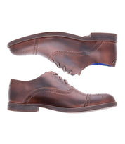 A pair of Bed|Stü Donatello II brown leather oxford shoes, crafted from premium vegetable-tanned leather, is shown from the side with one shoe upside down to display the classic cap-toe design, decorative stitching, and a slight heel.
