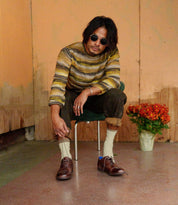 A man with medium-length hair and sunglasses sits on a chair indoors, wearing a striped sweater, rolled-up pants, cream socks, and Bed|Stü's Donatello II brown oxford shoes crafted from vegetable-tanned leather; a pot of orange flowers is beside him.