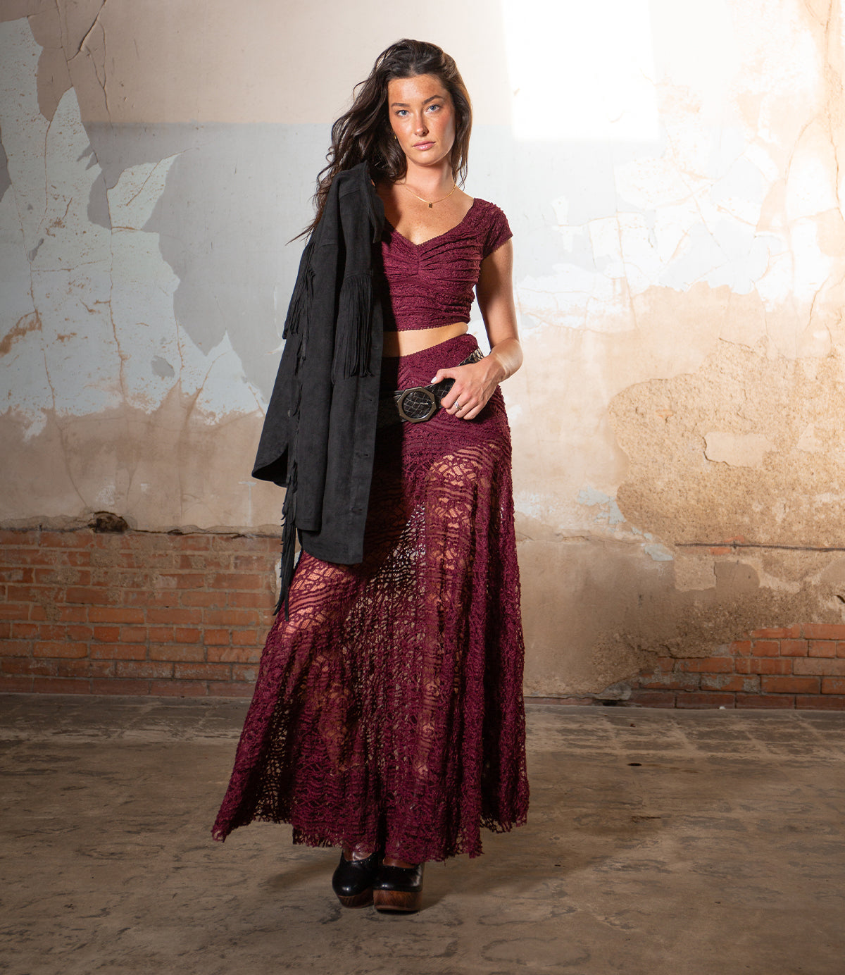 A woman wearing the Bed|Stü Dreamweaver maroon lace two-piece outfit and black platform shoes stands indoors against a worn, textured wall, holding a black jacket over her shoulder. Her look channels effortless bohemian flair, blending romantic textures from the Dreamweaver set with modern edge.