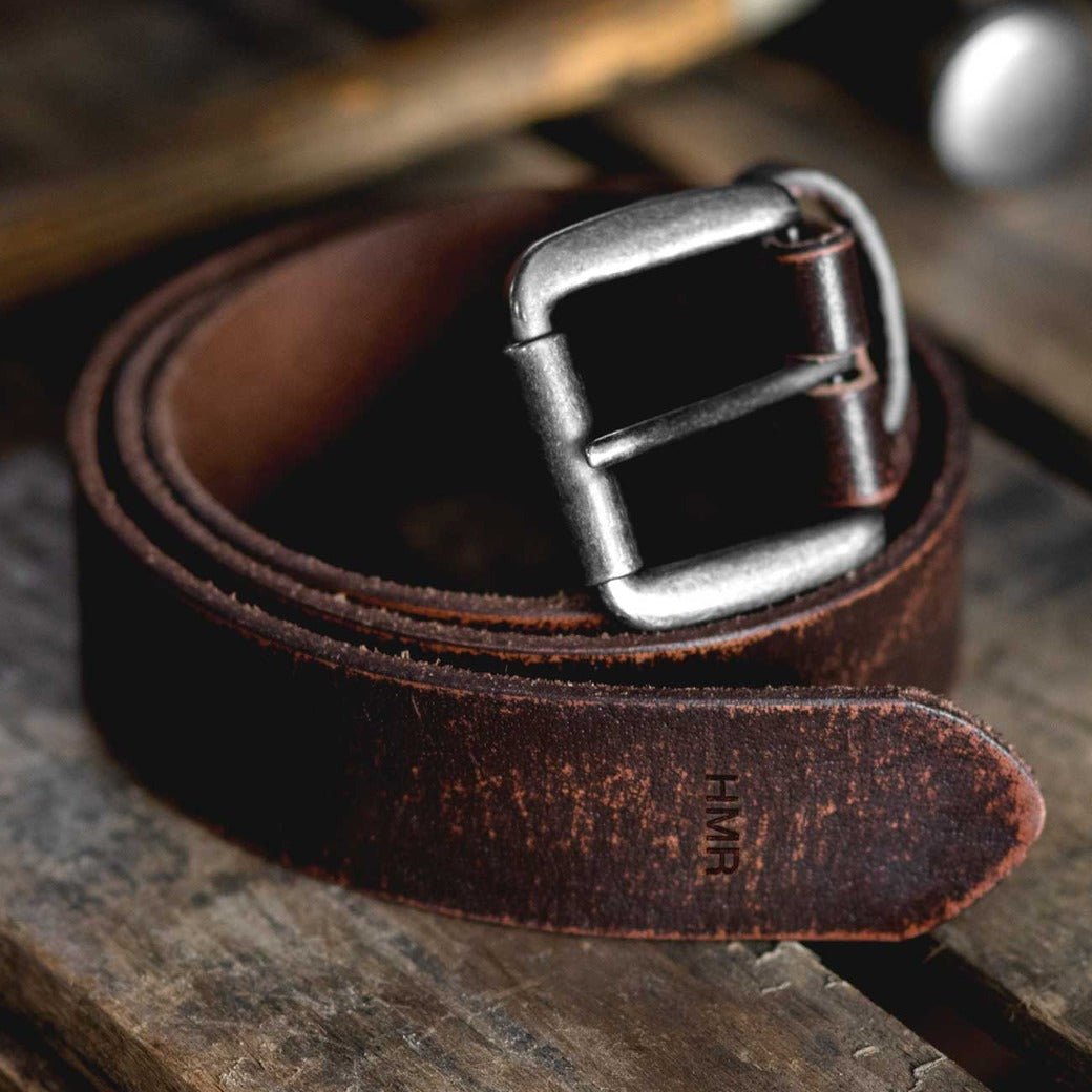 The Bed|Stü Drifter, a coiled brown leather belt featuring a metal buckle and textured leather strap, rests on a rustic wooden surface, embodying minimal style.