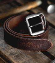 The Bed|Stü Drifter, a coiled brown leather belt featuring a metal buckle and textured leather strap, rests on a rustic wooden surface, embodying minimal style.