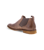Introducing the Bed|Stü Elude: a brown leather men's Chelsea boot featuring an elastic side panel, visible stitching, and a textured tan sole—all seen from a rear-side angle. This slip-on boot effortlessly merges classic style with everyday convenience.