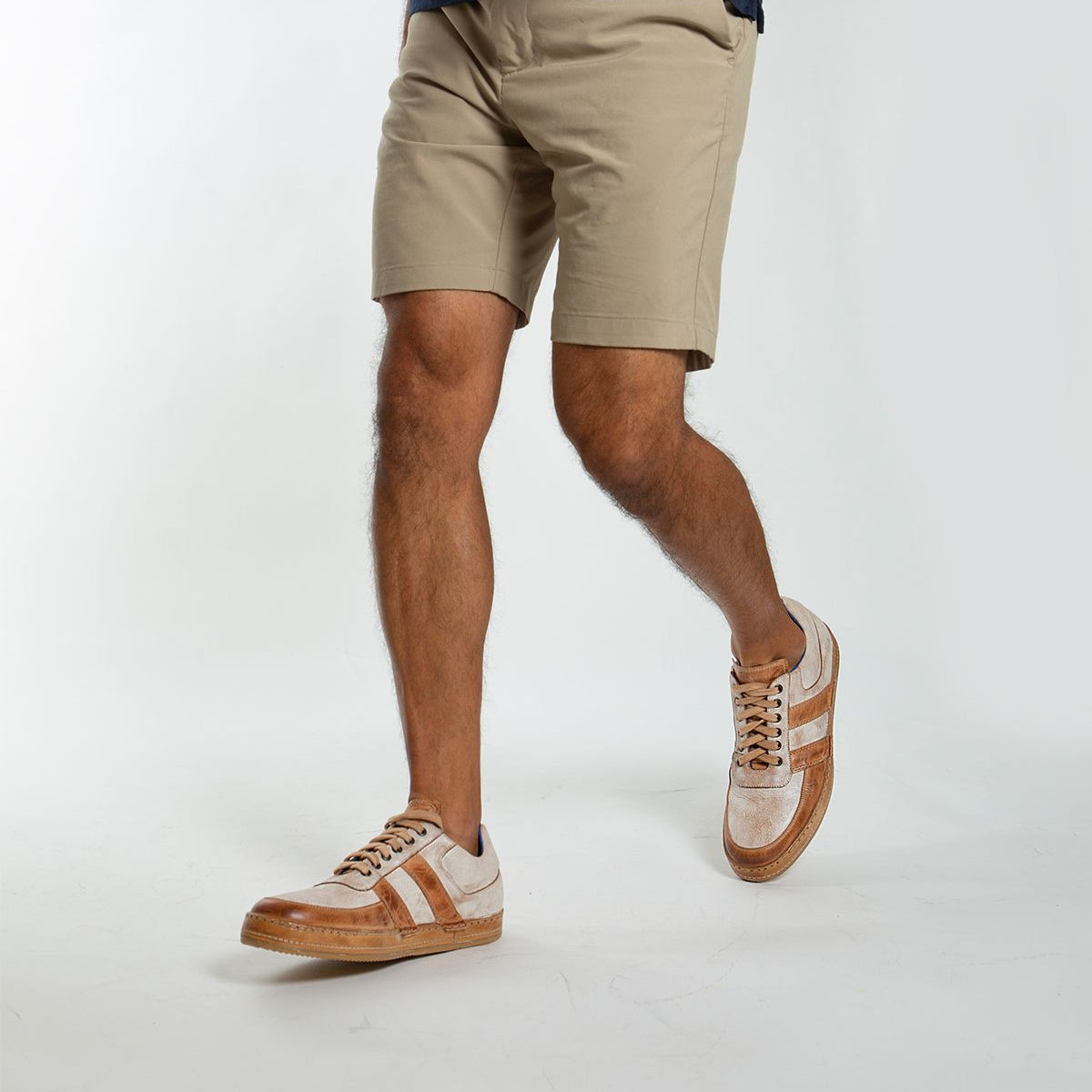 A man wearing beige shorts, a navy shirt, and Bed|Stü Firepower everyday sneakers—featuring leather stripe details and a textured sole—walks against a plain white background.