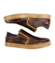 The Bed|Stü Freestyle features brown leather slip-on sneakers with yellow trim and tan soles, displayed in a side view with one shoe stacked on top of the other, providing a comfortable and stylish option for everyday wear.