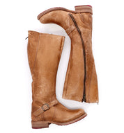 A pair of tall, brown leather Bed|Stü Glaye boots—one standing upright and the other on its side with the zipper partially open to reveal the interior—perfectly captures the classic equestrian boot style.