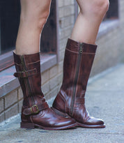 A person wearing the Bed|Stü Gogo Lug brown leather knee-high boots with zippers and buckles, featuring a sturdy lug outsole, stands on a sidewalk near a brick wall.