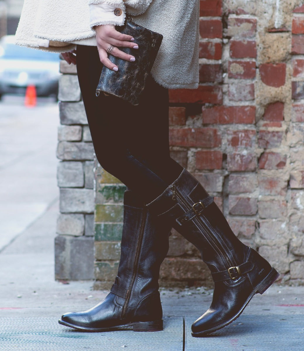 A person wearing Bed|Stü Gogo Lug Wide Calf boots and black leggings stands on a city sidewalk, holding a dark wallet, with a brick wall in the background.