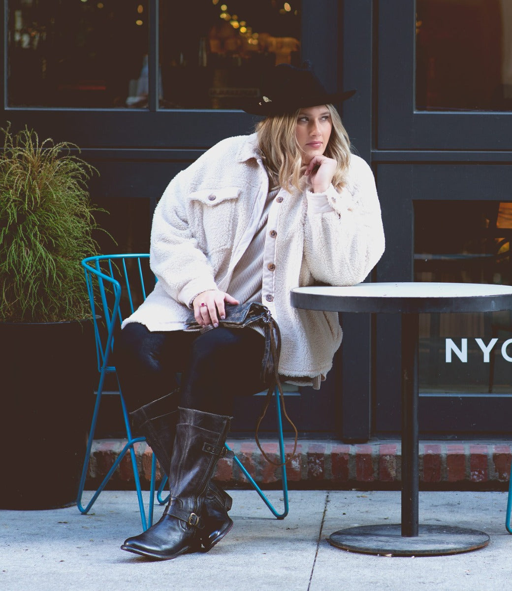 A woman wearing a black hat, cream jacket, and Bed|Stü Gogo Lug Wide Calf sleek leather mid-calf boots sits on a blue chair at an outdoor table, resting her elbow and looking to the side.