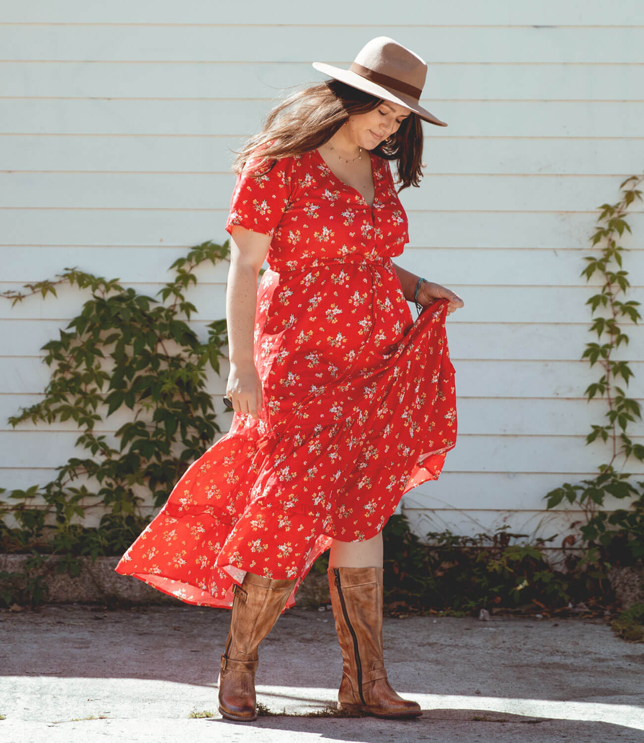 A woman stands outdoors near a white wall adorned with green vines, wearing a red floral dress, a tan hat, and Bed|Stü Gogo Lug Wide Calf leather boots while holding her dress.