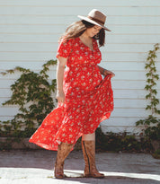 A woman stands outdoors near a white wall adorned with green vines, wearing a red floral dress, a tan hat, and Bed|Stü Gogo Lug Wide Calf leather boots while holding her dress.