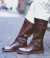 A person wearing the Bed|Stü Gogo Lug Wide Calf brown leather boots with buckles and zippers, standing on a paved sidewalk in light-colored pants.