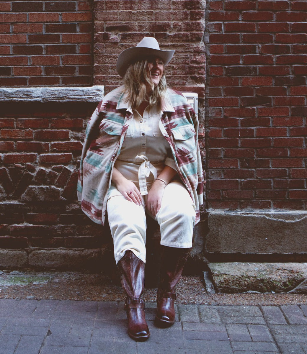 A person wearing a cowboy hat, a patterned jacket, cream shirt and pants, and Bed|Stü Gogo Lug Wide Calf boots sits smiling against an outdoor brick wall.