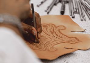 Craftsperson engraving a decorative pattern into a piece of tan leather by hand.