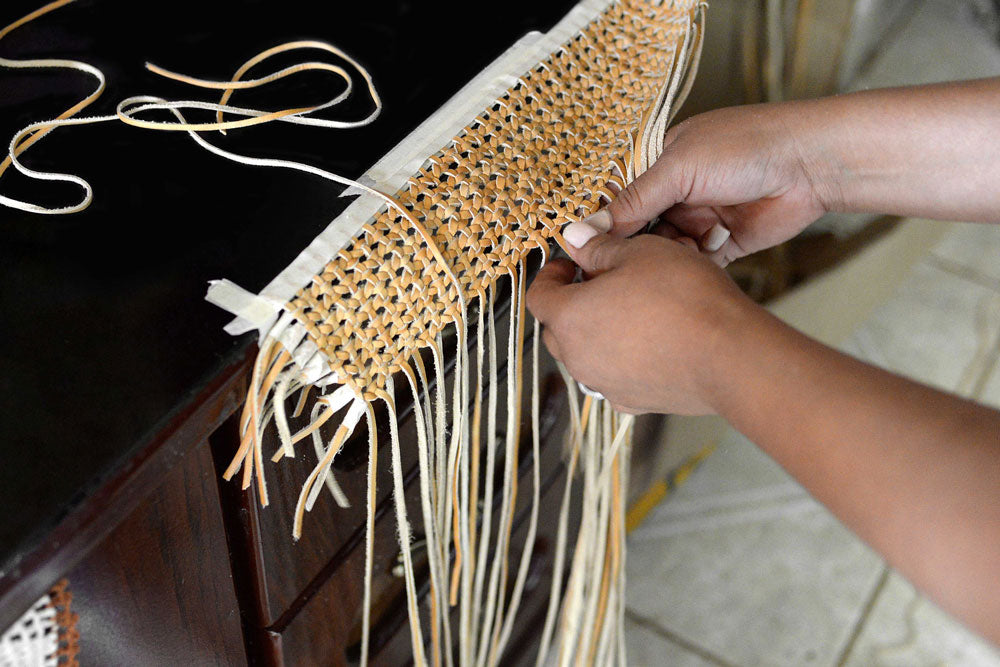 Hands weaving narrow strips of tan and cream leather into a patterned panel.