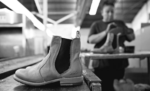 Black-and-white workshop photo of a Chelsea boot on a table, with a blurred craftsperson working on another boot in the background.