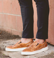 A person wearing the Bed|Stü Harry tan hand-dyed leather slip-on sneakers and black pants stands on a concrete surface next to a brick wall.