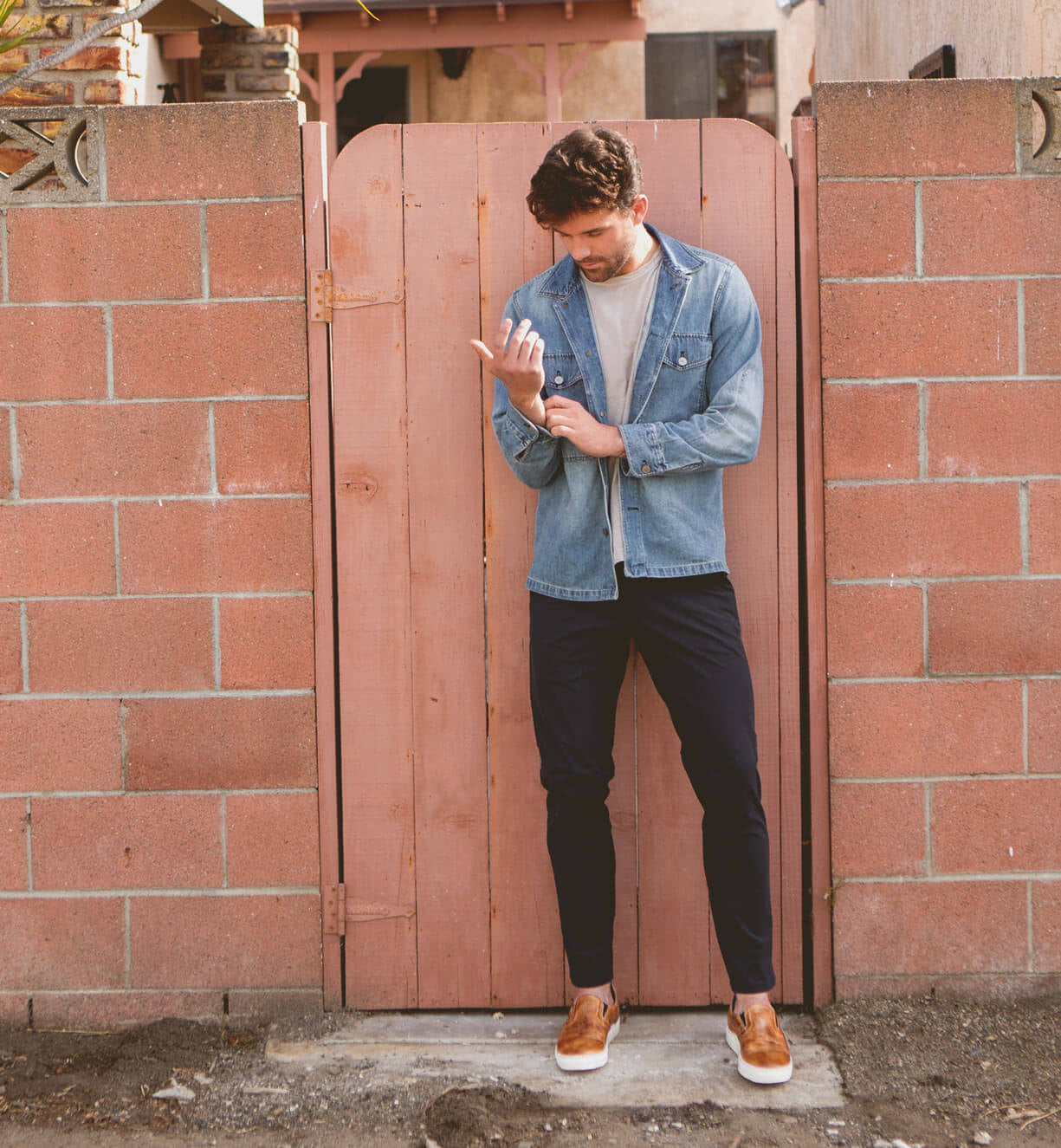 A man stands in front of a wooden gate and brick wall, wearing a denim jacket, white t-shirt, dark pants, and the Bed|Stü Harry slip-on sneakers featuring a lightweight outsole. He looks down while adjusting his sleeve.
