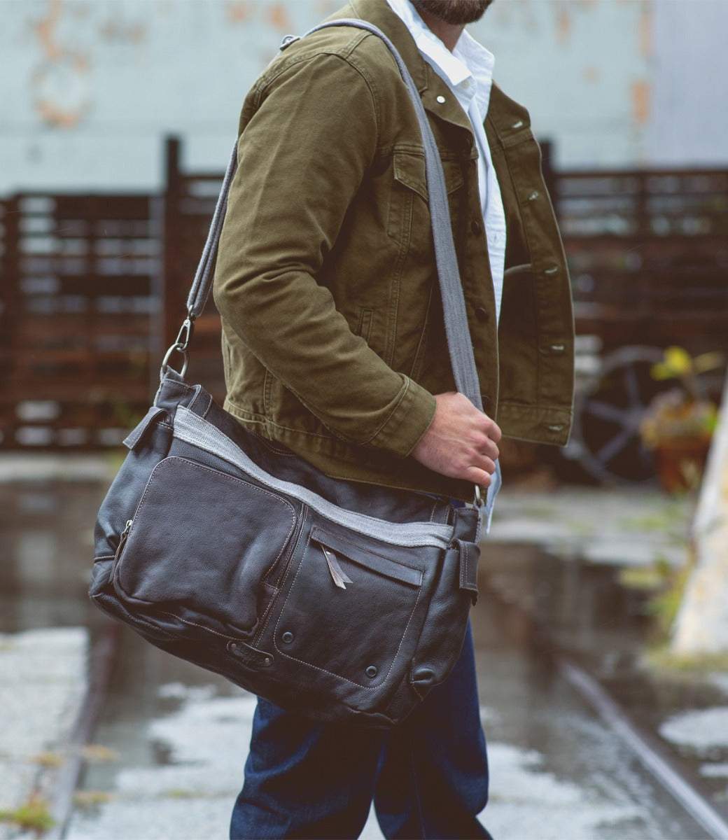 A man wearing a green jacket and white shirt carries the Bed|Stü Hawkeye II rugged leather messenger bag, featuring a padded laptop sleeve, over his shoulder while standing outdoors on a wet surface.