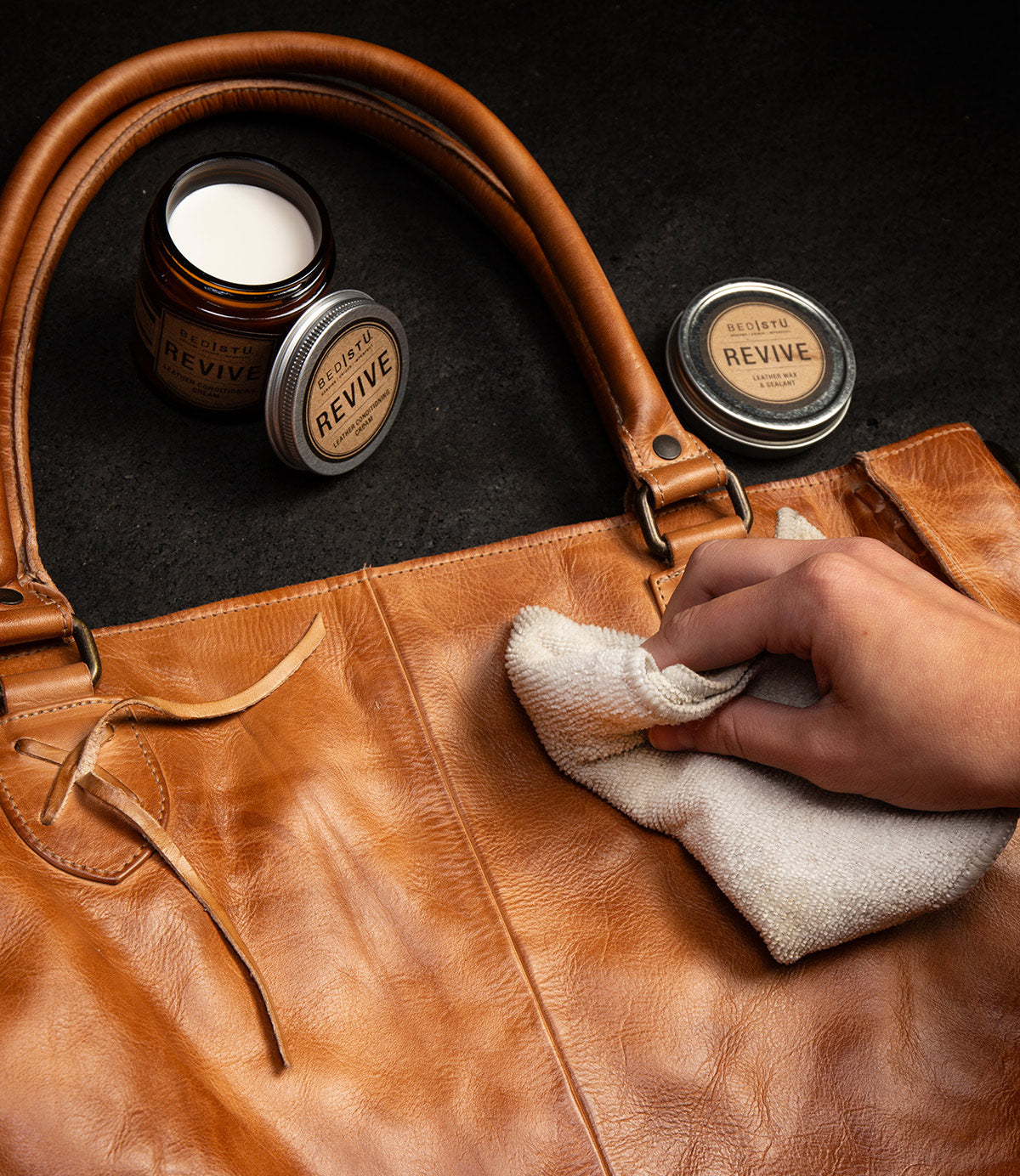 Hand polishing a tan leather bag with a cloth beside BED|STÜ Revive leather wax tins.