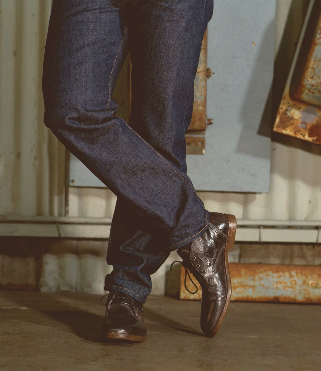 Standing indoors with legs crossed, the person is wearing dark blue jeans paired with Bed|Stü’s Illiad chukka shoes—polished black leather featuring a modern lace-up design. The background includes metal panels and a corrugated wall.