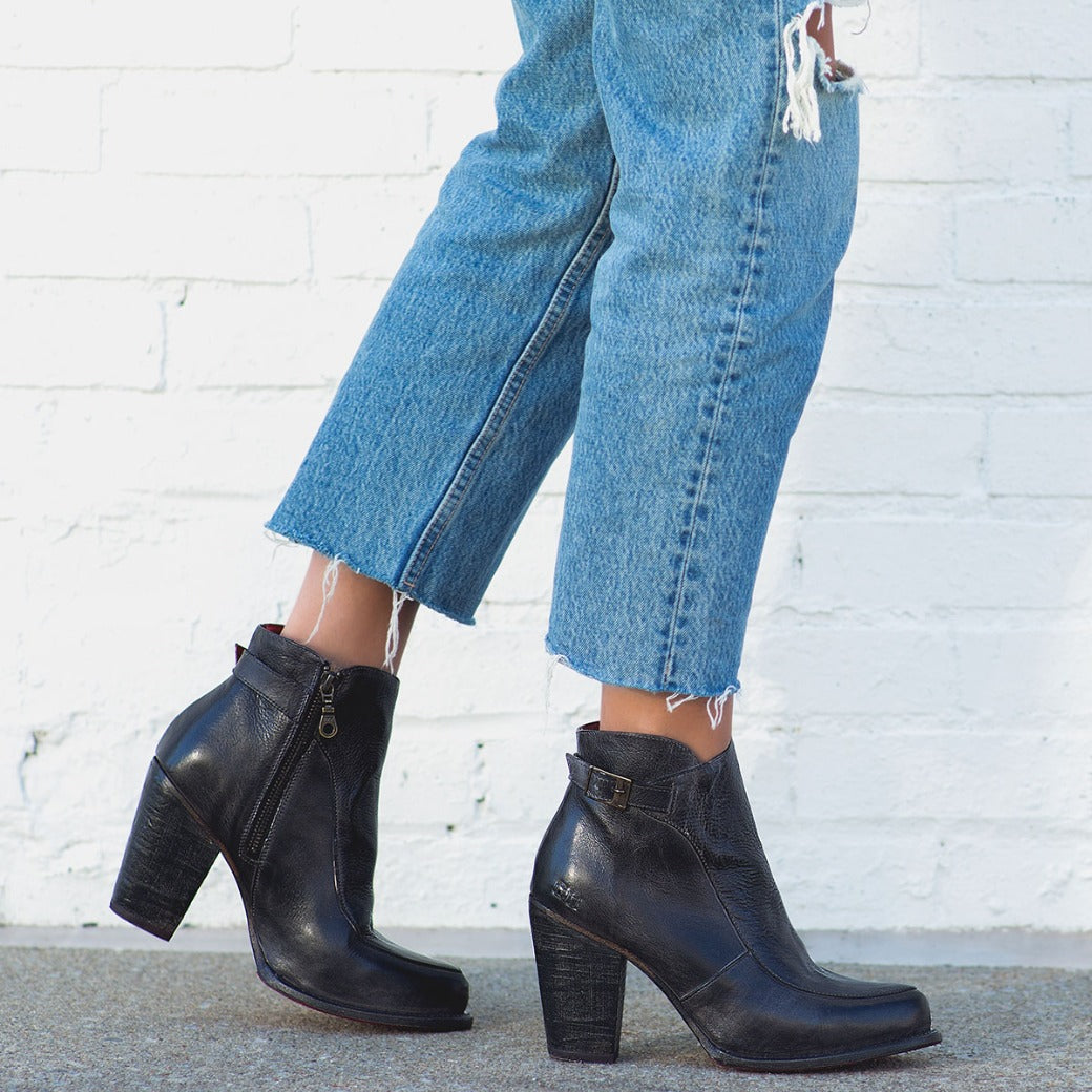 A person wearing the Bed|Stü Isla black heeled bootie and frayed blue jeans stands against a white brick wall on a concrete sidewalk.