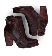 The Bed|Stü Isla booties are a pair of dark red smooth leather heeled shoes featuring chunky heels, side zippers, and decorative buckles, displayed on a white background.