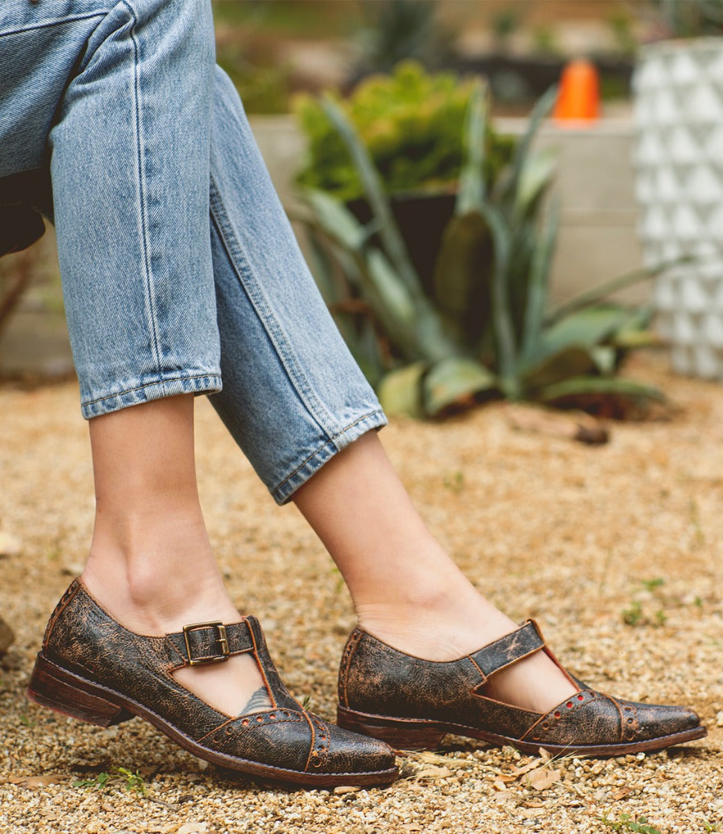 A person wearing light-wash jeans and the Bed|Stü Jenny vintage-style brown T-strap shoes with a low heel sits outdoors on a gravel surface with plants in the background.
