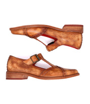 The Bed|Stü Jenny shoes are tan leather T-strap heels featuring a buckle closure, low heel, cushioned insole, and red inner lining, shown against a white background.