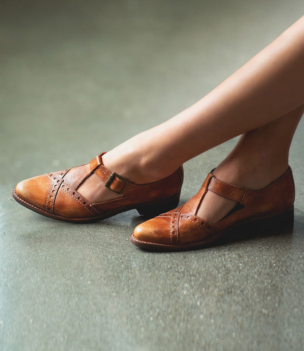 A person wearing Bed|Stü's Jenny brown leather T-strap Mary Jane shoes with cut-out details and a low heel, standing on a smooth gray floor.